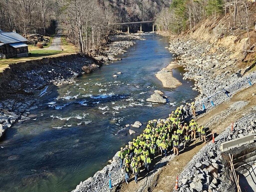 El equipo de Kiewit se reúne en la calzada reconstruida, restaurada después de que la tormenta arrasara más de 8.000 toneladas de escollera. A la izquierda, una propiedad vecina cercana a la obra lleva las cicatrices de la inundación, su ribera profundamente surcada por la ira de Helena.